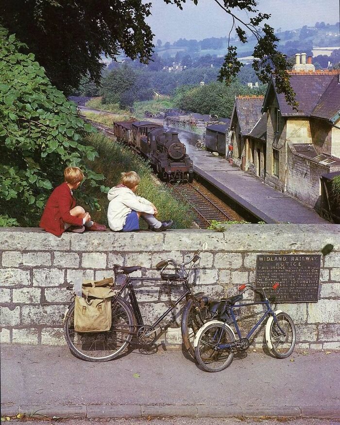 Children watching a vintage steam train pass by a rural station, with bicycles parked against a stone wall, evoking nostalgia.