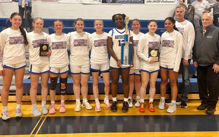 High school basketball team posing with their coach and trophies in a gymnasium.