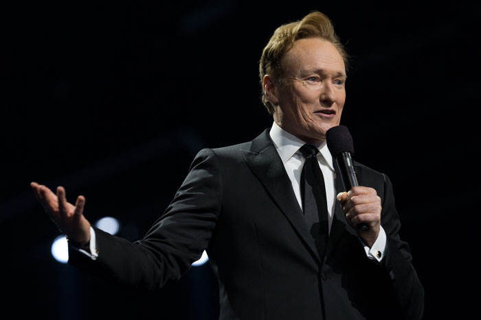 Man in a suit holding a microphone, delivering a monologue on stage with a spotlight backdrop, relating to controversial jokes at Oscars.