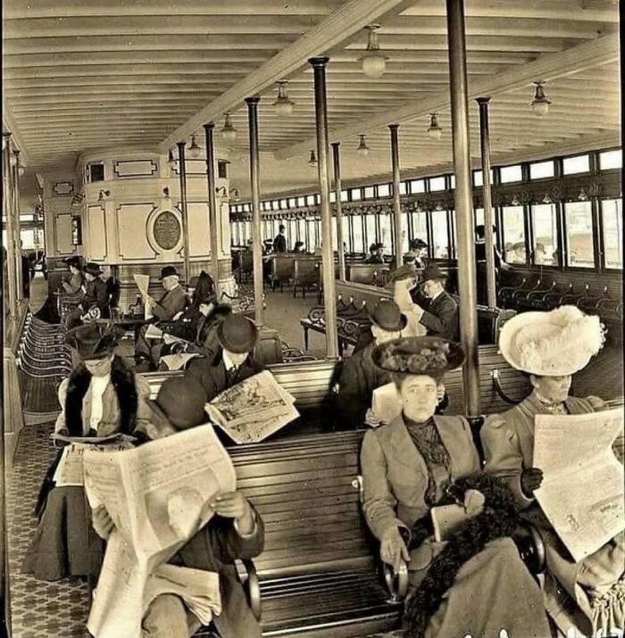 Historical photo of people in vintage attire reading newspapers on a ferry.