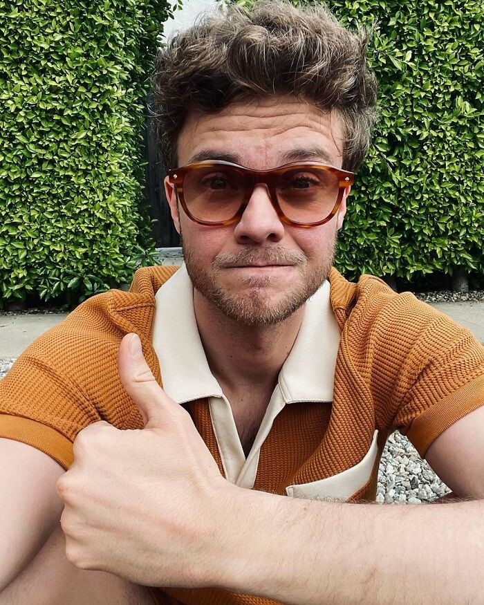 Young man in sunglasses and an orange shirt giving a thumbs up outdoors, symbolizing not-self-made celebs.