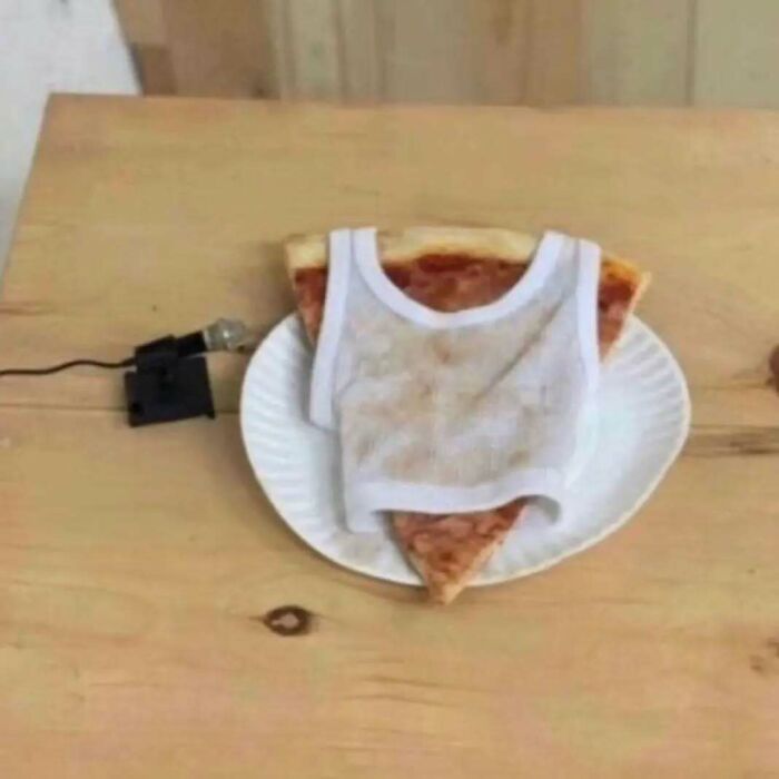 A slice of pizza on a plate, humorously dressed in a tiny white chaotic T-shirt, with a wire attached nearby.