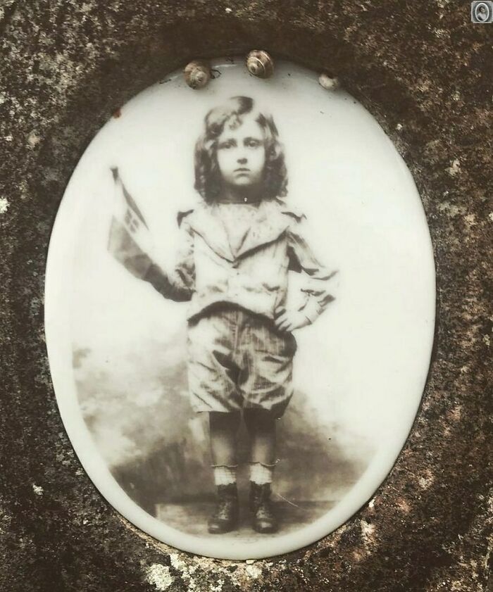 Vintage headstone portrait of a young child holding a small flag, set in an oval frame on a stone surface.