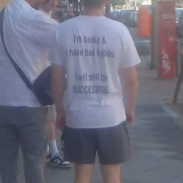 Man wearing a chaotic t-shirt with text about being broke and becoming successful, walking on a city street.