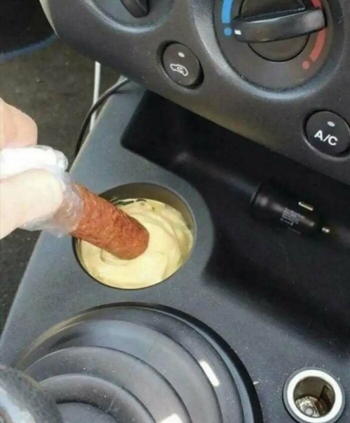 A sausage being dipped into mustard in a car cup holder, embodying food chaos.