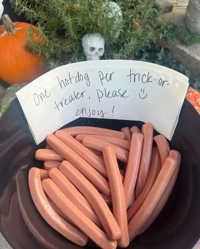 Hotdogs in a bowl with a sign reading "One hotdog per trick-or-treater, please enjoy!" amidst Halloween decorations.