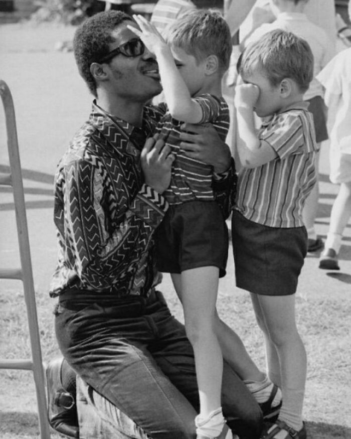 Man kneeling with two children in historical photo, wearing sunglasses and patterned shirt, sharing a joyful moment outdoors.