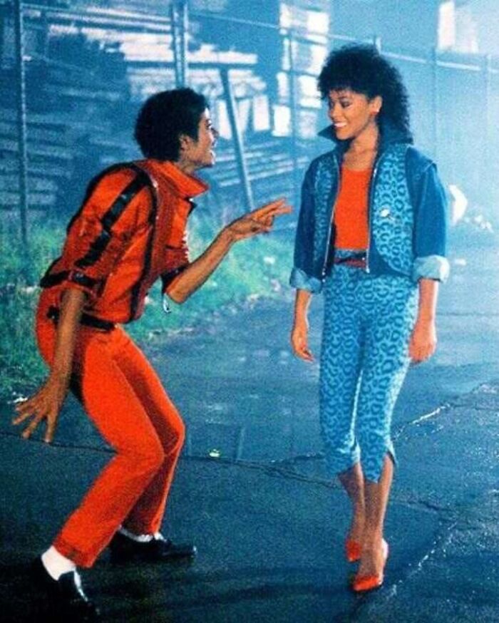 A man in a red outfit enthusiastically gestures to a woman wearing blue and red, evoking historical pop culture.