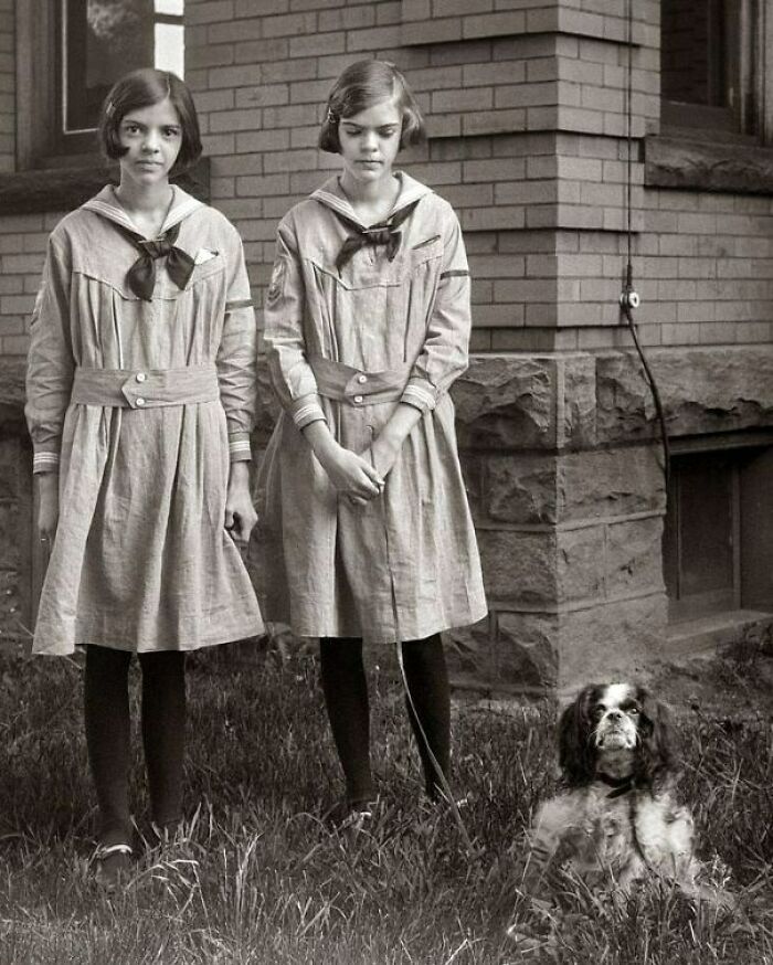 Two girls in vintage dresses with a dog, standing in front of a brick building, reflecting historical moments of the past.