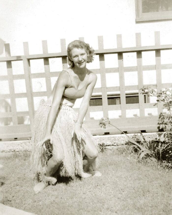 A woman in a grass skirt poses with a smile in a vintage garden setting, showcasing a historical moment.