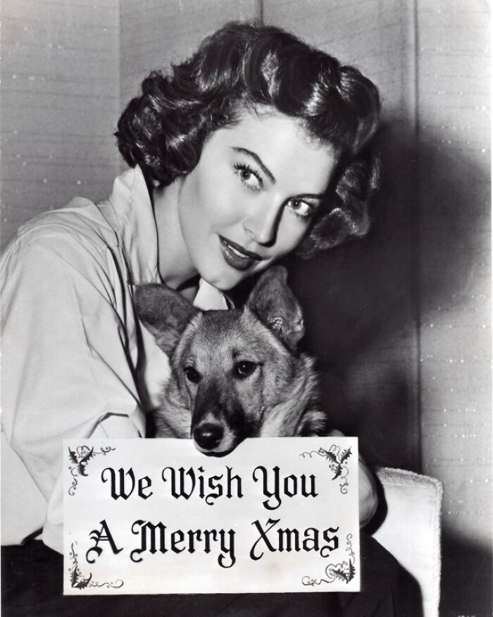 Woman with vintage hairstyle and a dog holding a "We Wish You A Merry Xmas" sign, reflecting historical charm.