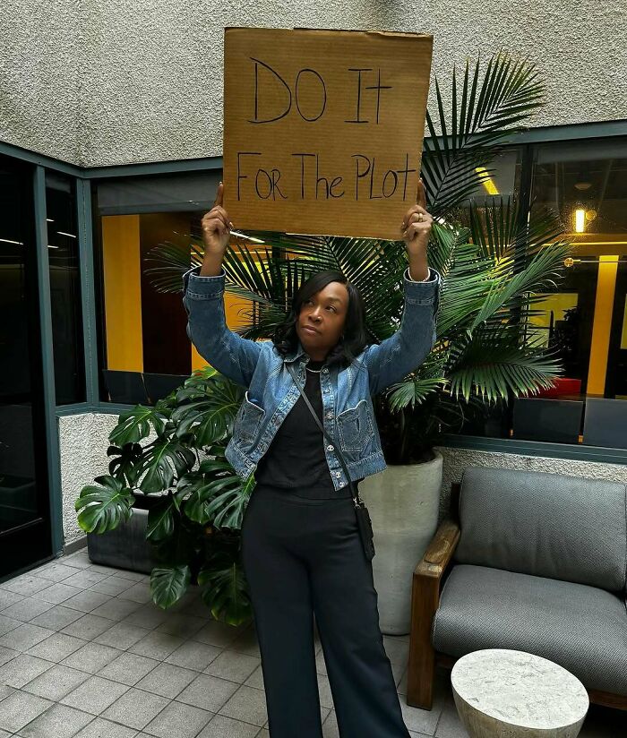 Person holding a sign that says "Do it for the plot" in a courtyard with plants, capturing a petty moment.