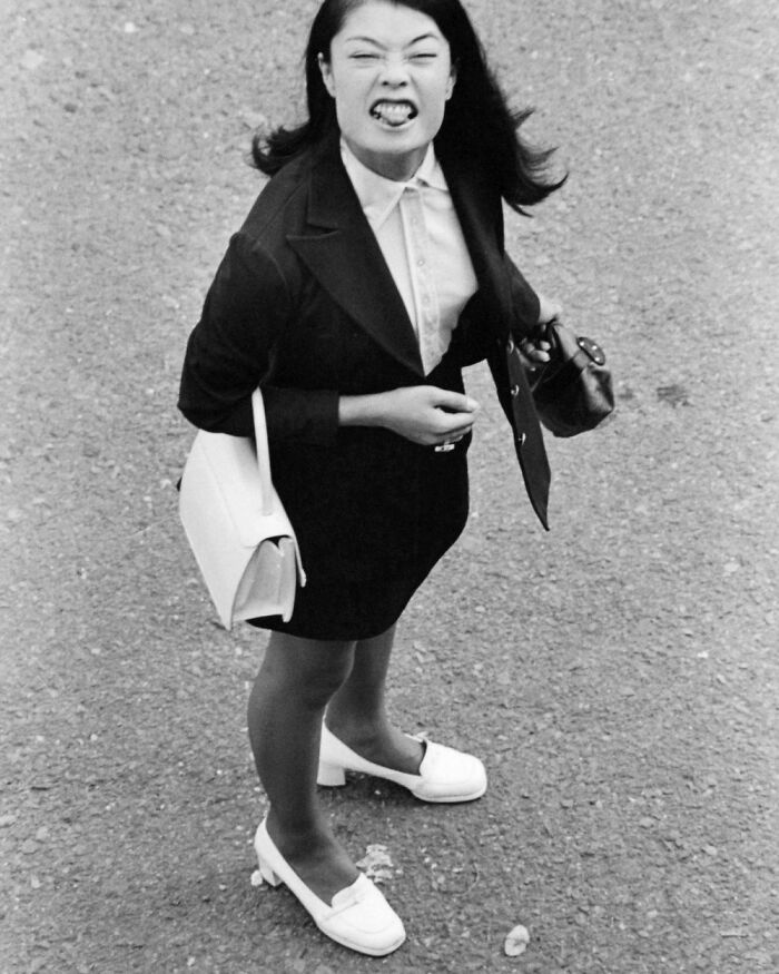 Historical photo of a woman in a suit and white shoes making a face, holding a purse and bag, standing on a street.
