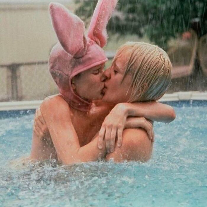 Two people embracing in a pool, one wearing a pink bunny hat, capturing a wild and wholesome '90s moment.