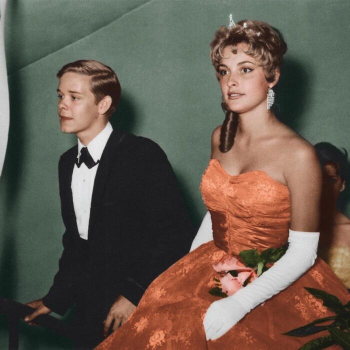 Two young people in formal 1950s attire at a prom; woman in an orange gown, holding flowers. Historical fashion insight.