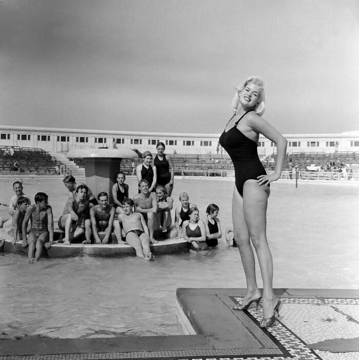 Woman in vintage swimsuit posing by a pool, surrounded by people, offering a historical glimpse into past fashion trends.