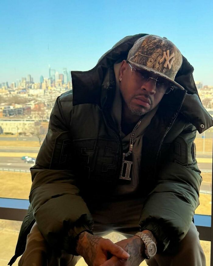 Man in camouflage cap and dark jacket with cityscape, highlighting celebrity lifestyle choices.