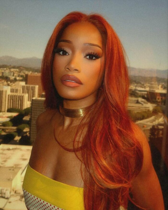 Celebrity with red hair and bold makeup stands against a cityscape backdrop, embodying a petty celebrity moment.