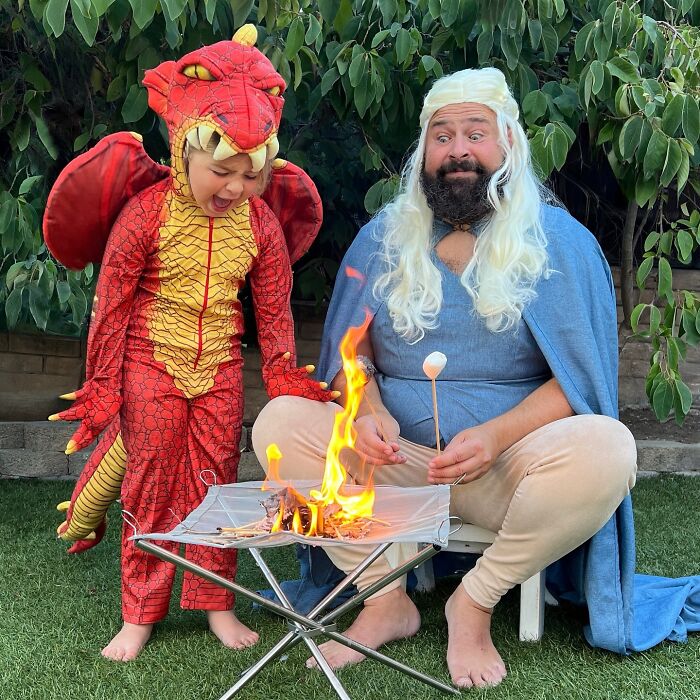 Dad and daughter in costumes, dad with a marshmallow at a pretend campfire, daughter dressed as a dragon.
