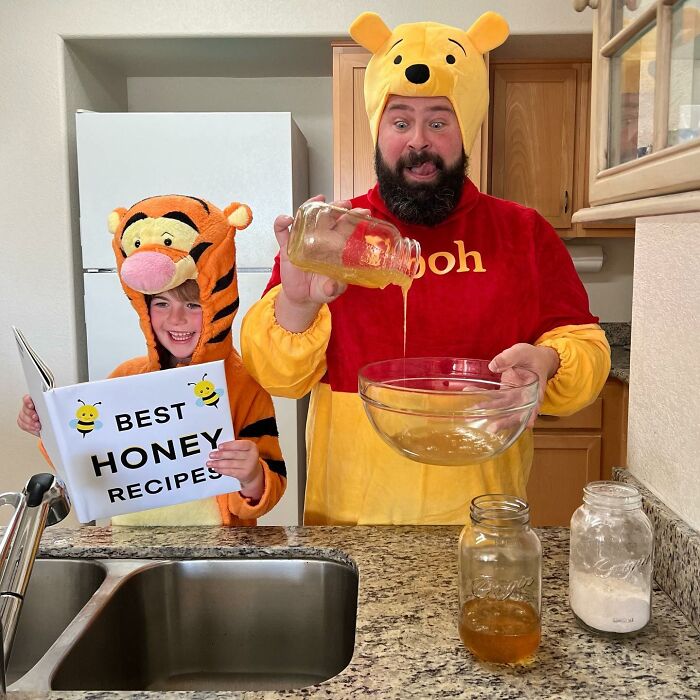 Dad and daughter in costumes, dad as Winnie the Pooh, daughter reading "Best Honey Recipes" book, having fun in kitchen.