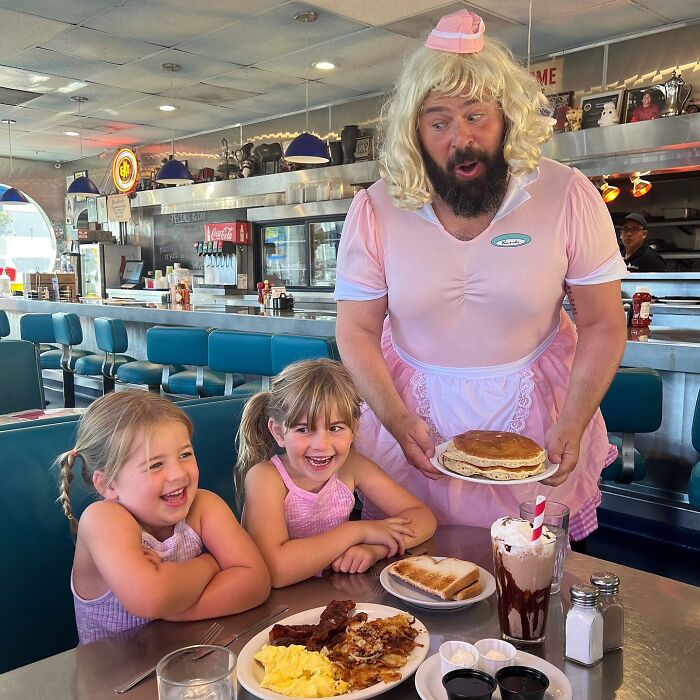 Dad in costume serves pancakes to smiling daughters in a retro diner.