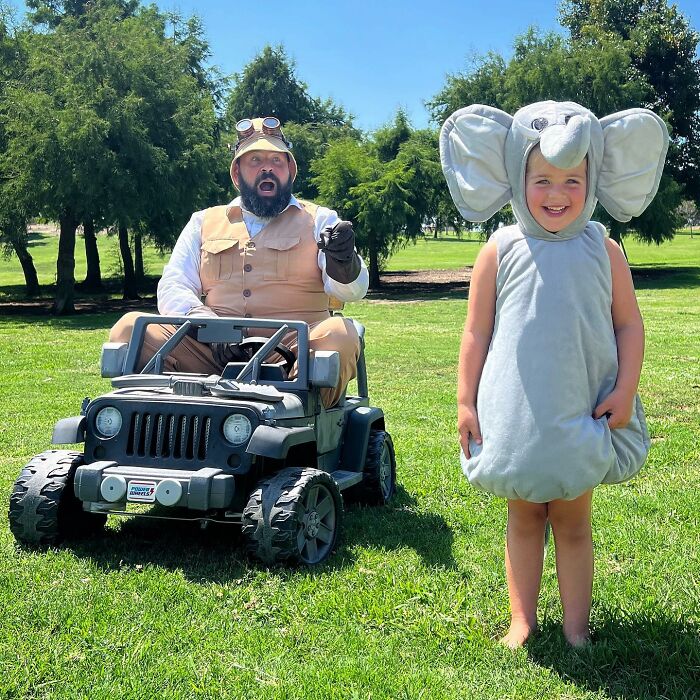 Dad in adventurer outfit in toy jeep with daughter in elephant costume, having fun outdoors.