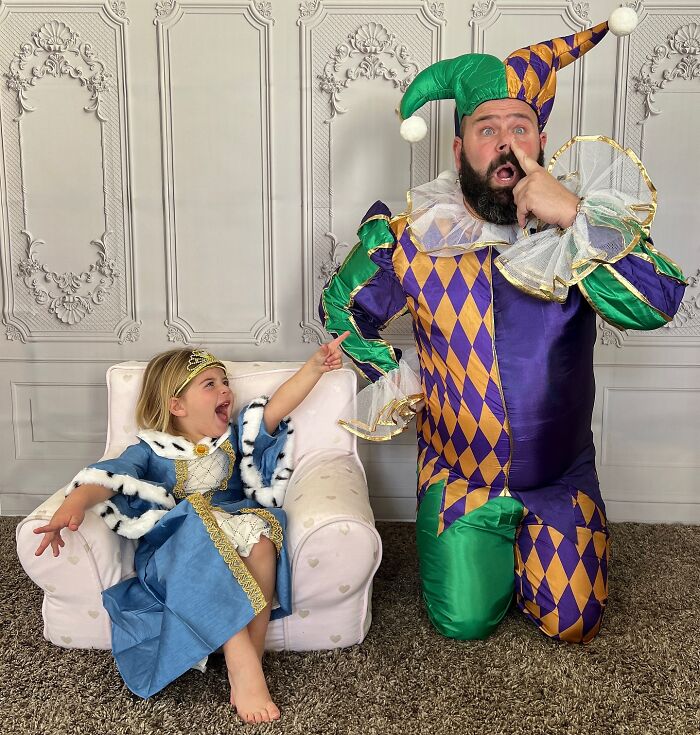Dad and daughter in funny costumes; he’s dressed as a jester and she as a princess, both laughing together.