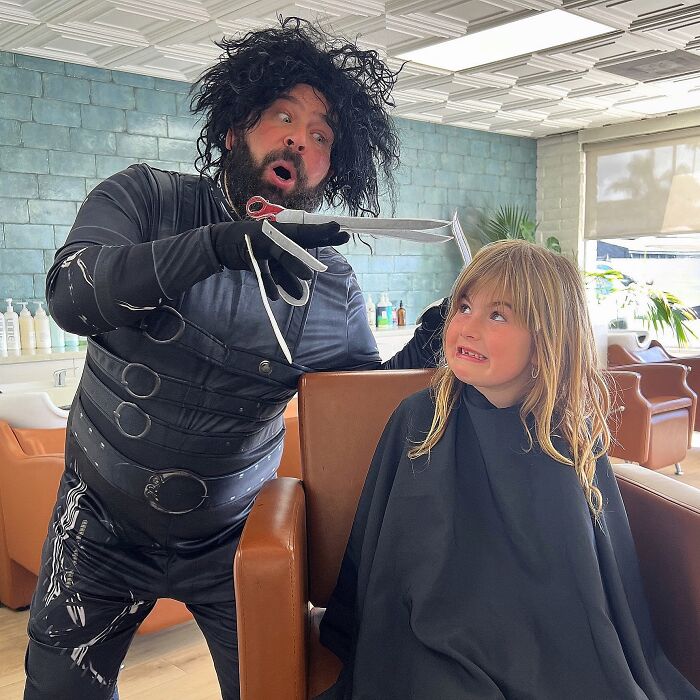 Dad in funny costume mimics haircutting with daughter at salon, both making amusing expressions.