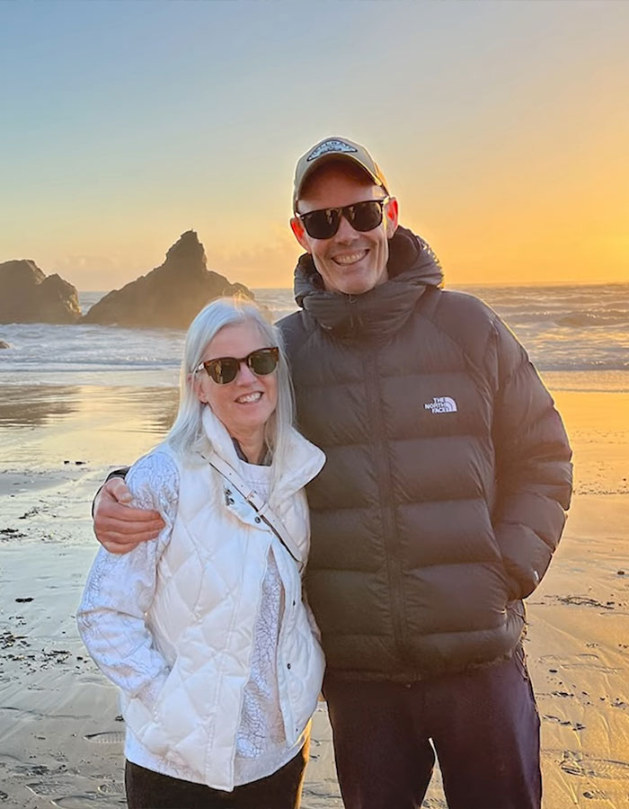 A couple in sunglasses smiling on a beach at sunset.