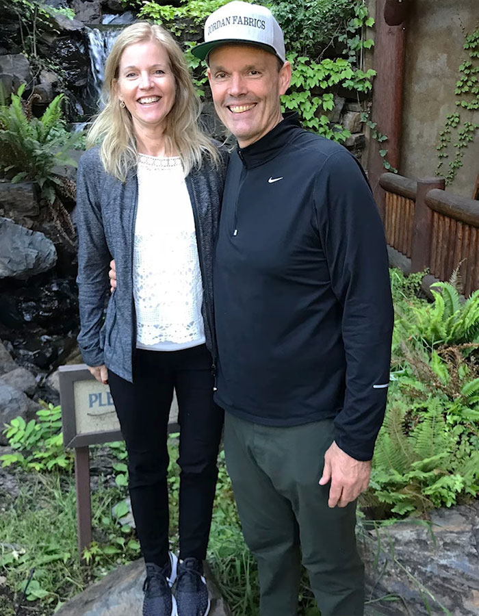 A couple smiling outdoors near foliage and a small waterfall.