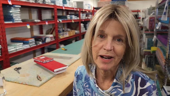 Beloved YouTuber sits in a craft room filled with supplies, wearing a patterned shirt, speaking to her audience.