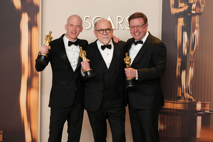 Three winners from the Oscars 2025 holding statuettes, dressed in tuxedos and standing before an Oscars backdrop.