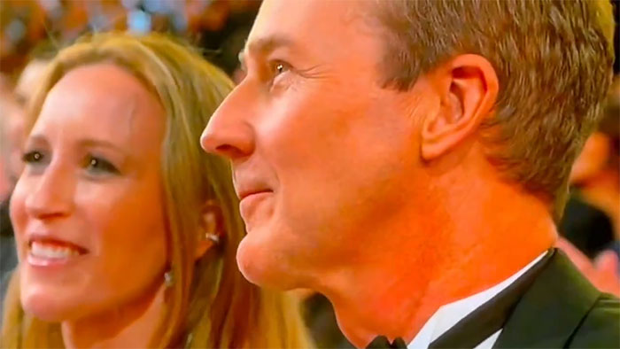 Man in tuxedo at an award ceremony, reacting to a winner announcement, woman smiling beside him; capturing expressive moment.