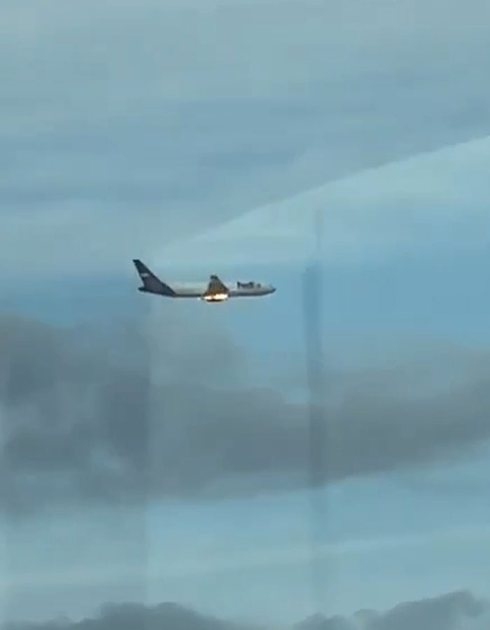 FedEx aircraft ablaze mid-air causing packages delayed, against a backdrop of cloudy sky.