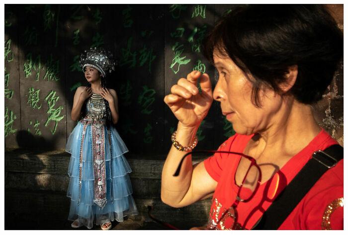 A woman adjusts her glasses in foreground, while another in traditional attire poses with green wall. Street photography by Andrés Ramos.