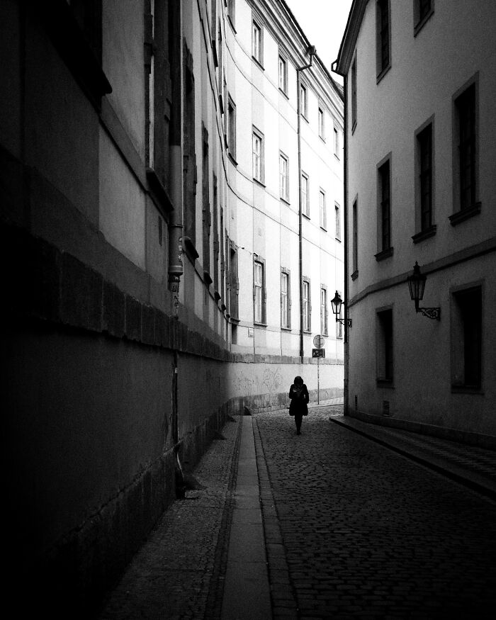 Black-and-white street photograph of a solitary figure walking down a narrow alley between tall buildings.