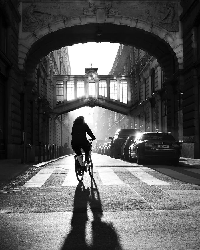 Black-and-white street photograph of a person biking under an ornate archway, casting a long shadow.