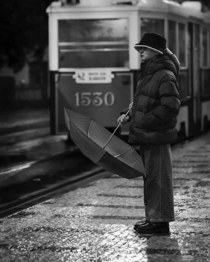 A person with an umbrella stands on a wet street near a tram in a black-and-white street photograph.