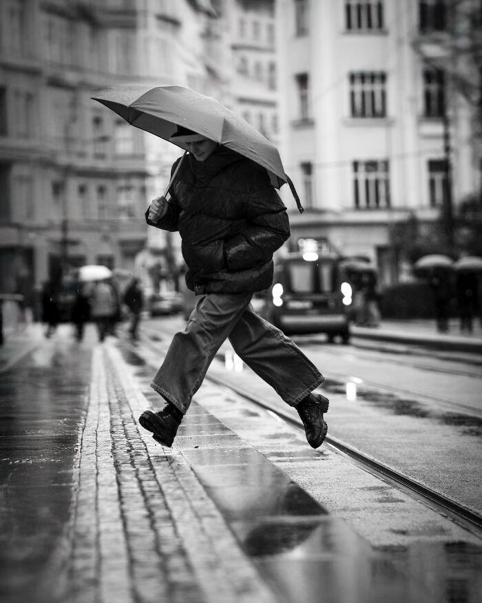 Child with umbrella leaping over puddle in stunning black-and-white street photograph.