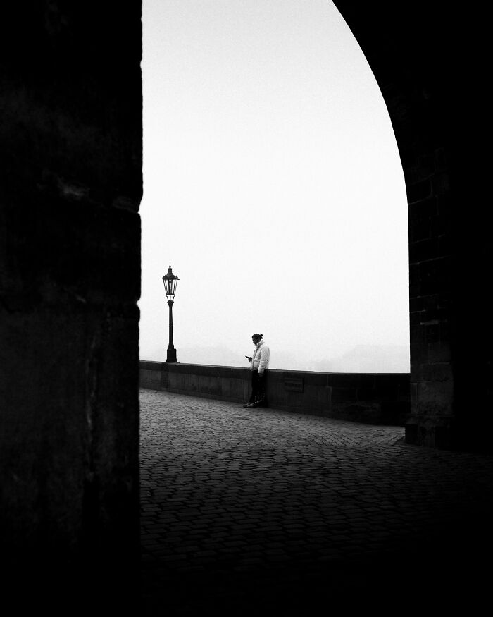 Black-and-white street photograph featuring a person standing under an archway beside a lamppost.