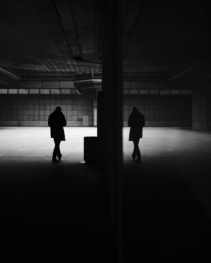 Silhouetted figure walking in an urban black-and-white street scene, casting a reflection on a wall.