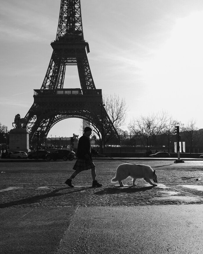 A person walking a dog near the Eiffel Tower in a black-and-white street photograph.
