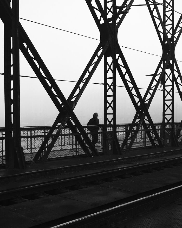 Black-and-white street photograph of a person walking on a foggy bridge, framed by metal beams.