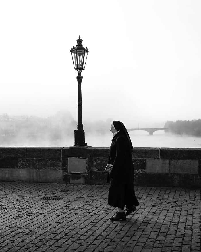 Nun walking on cobblestone street by river; black-and-white street photograph with lamppost and misty bridge in background.