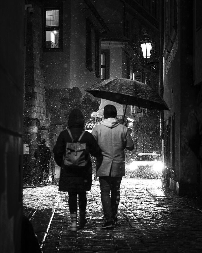 A couple under an umbrella walks through a cobblestone street at night, capturing black-and-white street photography charm.