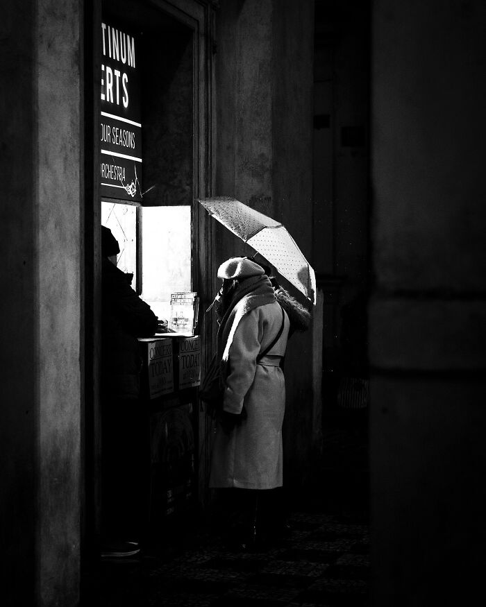 A person with an umbrella stands at a ticket booth in a black-and-white street photograph.