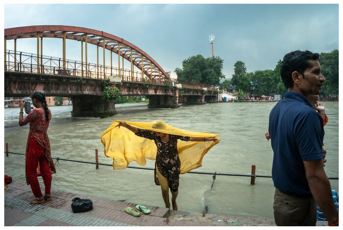 A person drying a yellow cloth by a river, with a bridge in the background, capturing a raw street moment.