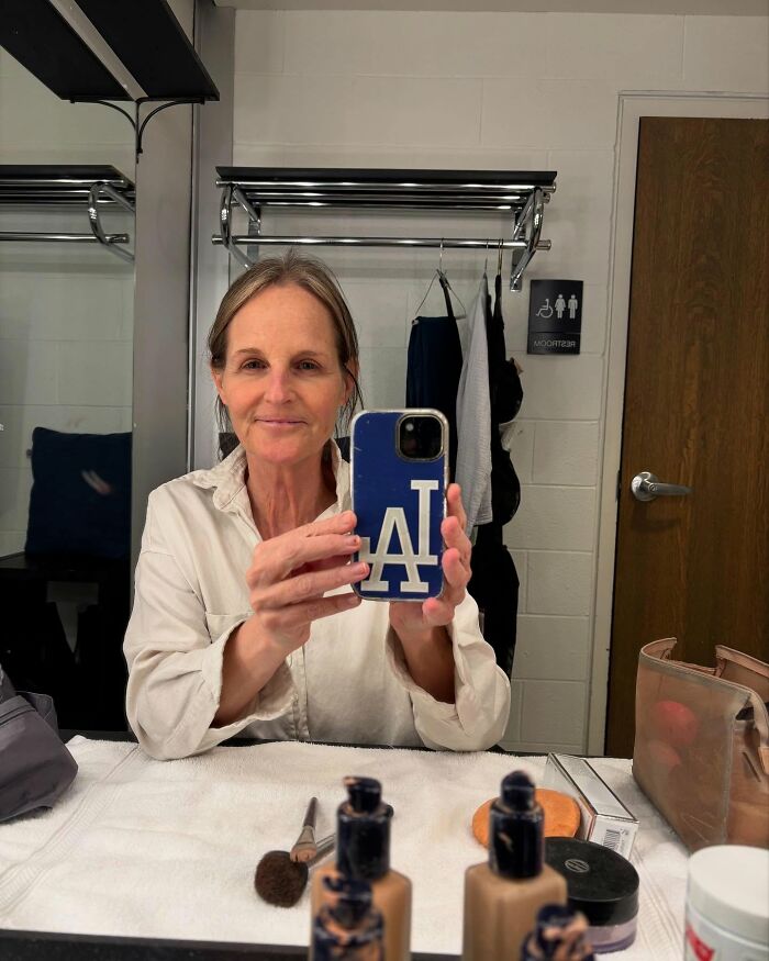 A woman takes a selfie in a dressing room, holding a phone with an LA logo, wearing a white shirt. A woman takes a selfie in a dressing room, holding a phone with an LA logo, wearing a white shirt.