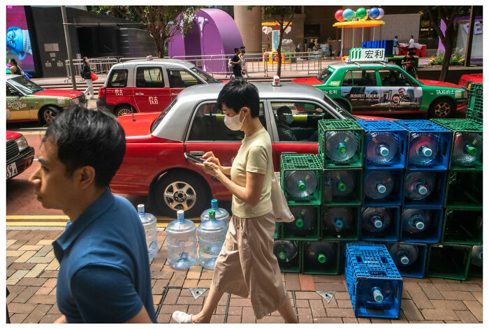Street scene with people walking beside stacked water bottles in front of colorful taxis, capturing raw urban moments.