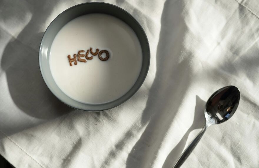 A bowl of cereal spelling "Hello" in milk, with a spoon on a white cloth.